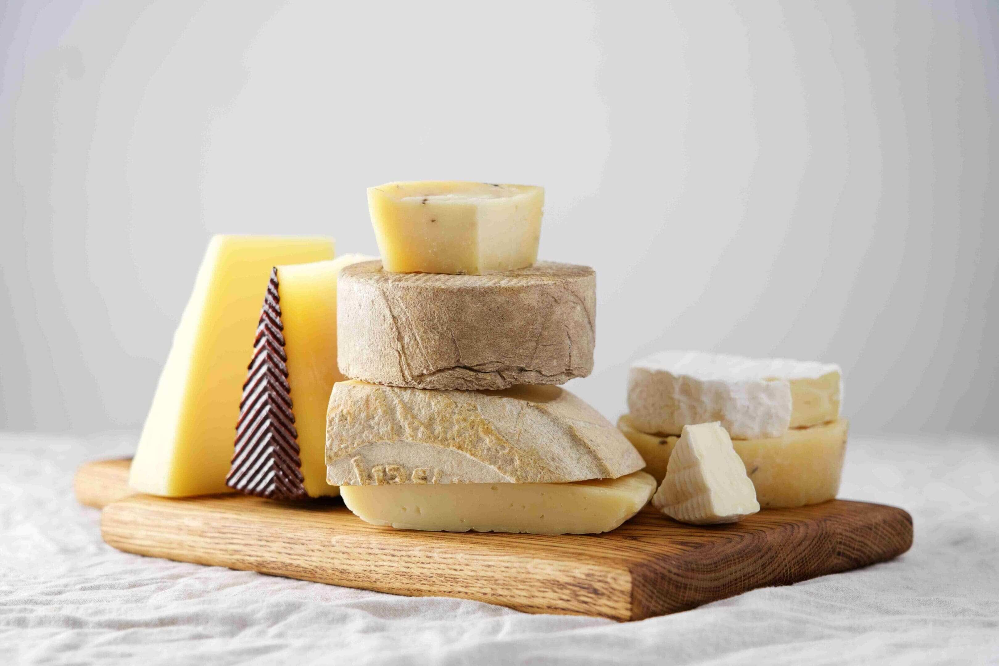 Assorted cheeses stacked on a wooden board against a neutral background.